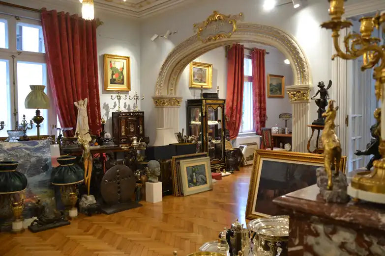 O cameră plină cu diverse mobilier antic, picturi, sculpturi și obiecte decorative, probabil pentru vânzare.